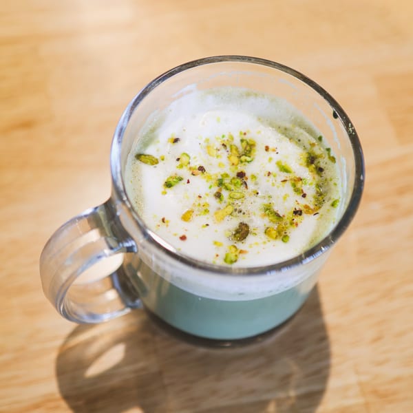A glass mug of green chai latte with foamed milk and pistachio sprinkles on a wooden table.