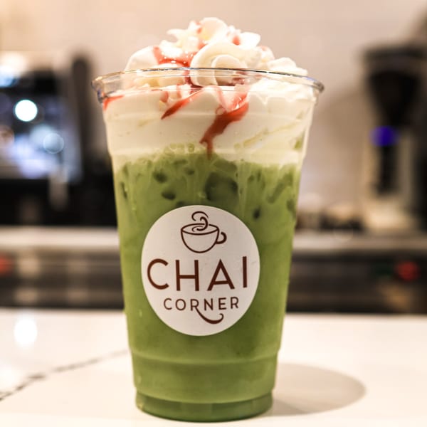 Iced green drink with whipped cream and red drizzle in a Chai Corner branded cup.