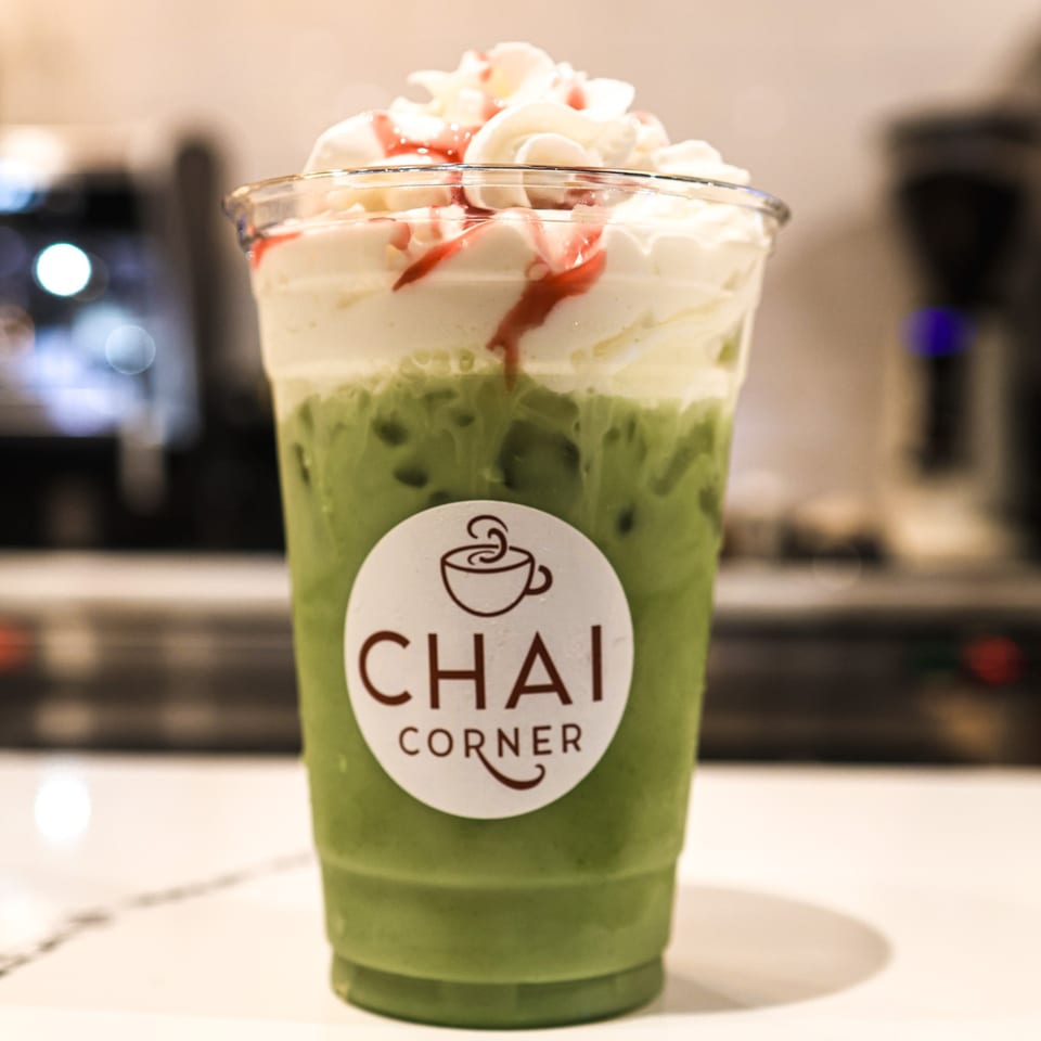Iced green drink with whipped cream and red drizzle in a Chai Corner branded cup.