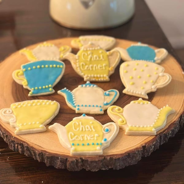 Chai Corner cookies shaped like teacups and teapots with yellow and blue icing on a wooden board.