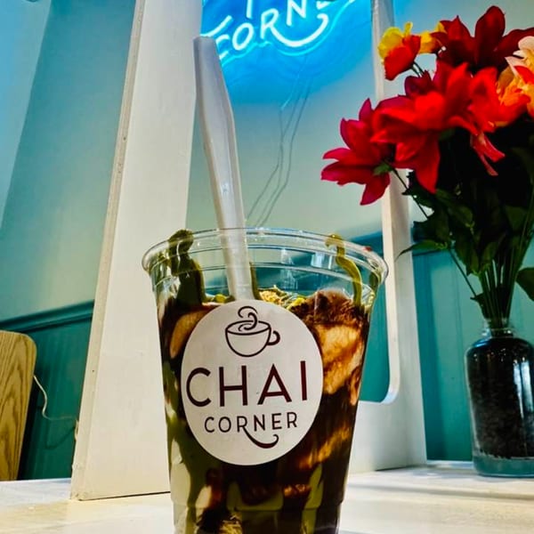 Chai Corner dessert cup with chocolate drizzle and a spoon, next to red flowers and a neon sign.